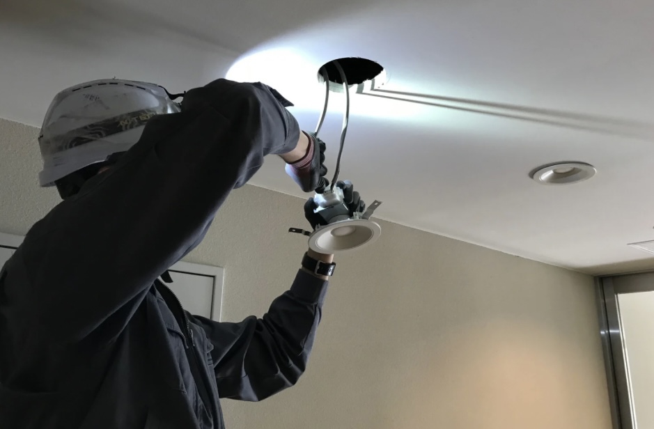A professional electrician installing a recessed light fixture in a ceiling, using tools and wearing safety gear.